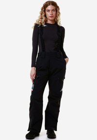Pantaloni da sci neri impermeabili con bretelle regolabili, tasche laterali con zip e dettagli del logo, indossati sopra una maglietta a maniche lunghe di colore scuro.
