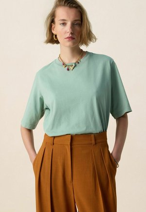 Jeune femme aux cheveux blonds courts portant un t-shirt vert menthe, un pantalon plissé taille haute couleur rouille, des boucles d'oreilles anneaux dorés et un collier avec un pendentif.