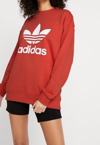 Röd sweatshirt i mjuk bomullsblandning, med en stor vit Adidas-logotyp och tre-stripesdesign på ärmarna. Parat med svarta shorts.