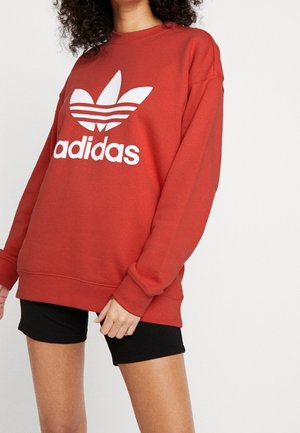 Un sweat-shirt rouge en mélange de coton doux, arborant un grand logo Adidas blanc et un design à trois rayures sur les manches. Associé à un short noir.