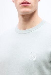 Gros plan sur une personne portant un pull gris clair à col rond avec un petit patch logo "BOSS" sur la poitrine.