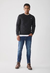 Zwart sweatshirt met een ronde hals en witte accenten, gecombineerd met een blauwe spijkerbroek en bruine veterlaarzen, met een klein logo op de borst.