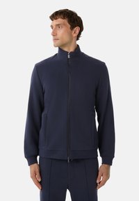 Mand iført en marineblå zip-up jakke og matchende bukser, står med hænderne afslappet og kigger til venstre mod en ensfarvet baggrund.