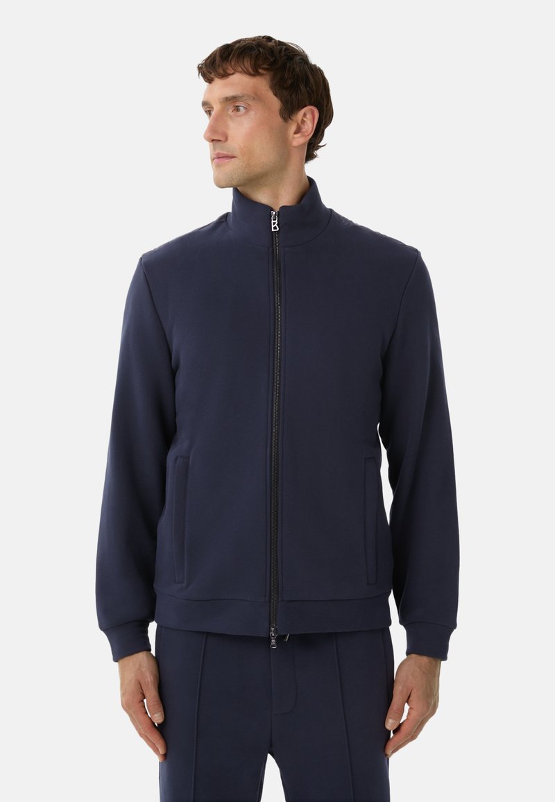 Mand iført en marineblå zip-up jakke og matchende bukser, står med hænderne afslappet og kigger til venstre mod en ensfarvet baggrund.