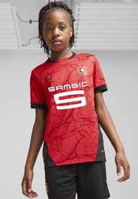 Raudona futbolo marškinėliai su juodais akcentais, turintys geometrišką raštą ir didelį baltą tekstą "SAMSIC 6". Juodi šortai papildomi drabužį.