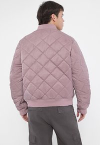 Quilted bomber jacket in mauve, featuring a ribbed neckline and cuffs, with textured fabric. Worn with gray cargo pants.