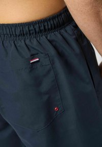 Temno mornarsko modre kratke hlače z elastičnim pasom, z zadnjim žepom, na katerem je majhen rdeče, belo in modro obarvan logotip. Gladek tekstura in priložnostna zasnova.