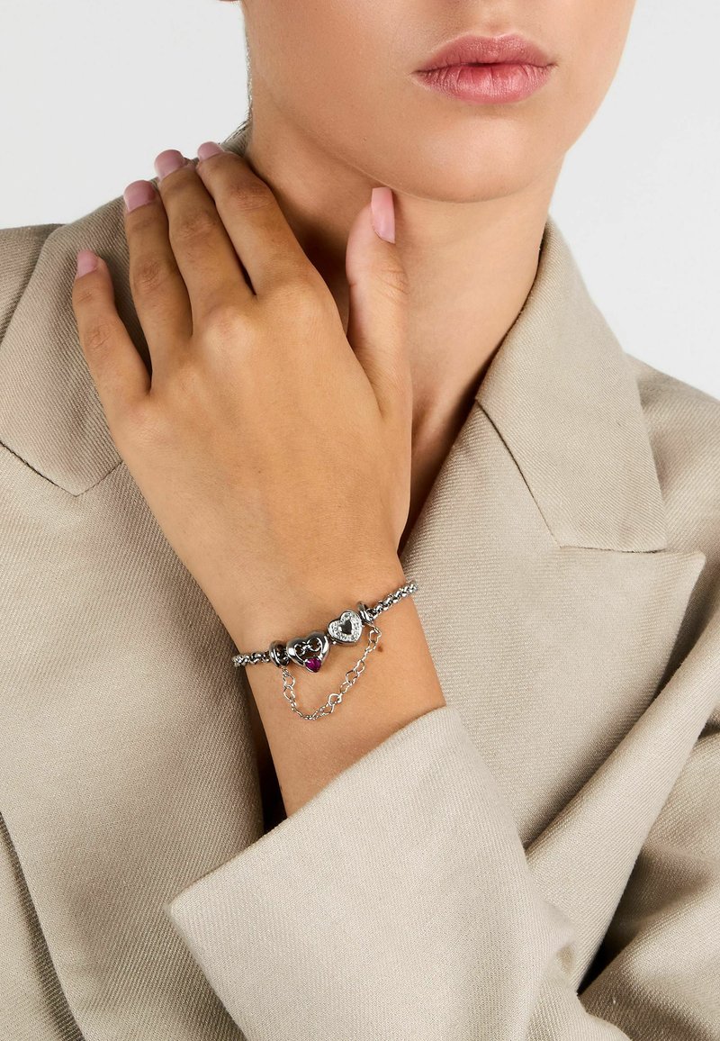 Bracciale in argento a catena con charm a forma di cuore, uno con un accento rosa e l'altro con dettagli decorativi, su una mano contro uno sfondo beige.