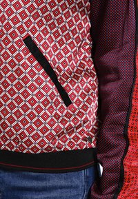 Red and white patterned jacket featuring a diamond design, with a black zipper pocket and contrasting textured sleeve in dark red.
