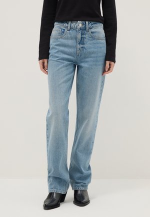 Vrouw die lichtblauwe, rechte, hoogwater spijkerbroek draagt, een zwart lang mouwen shirt en zwarte laarzen met blokhak en vierkante neus, staand op een witte vloer.