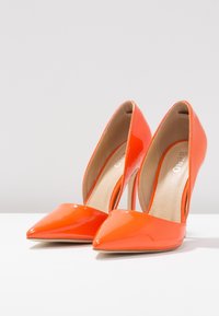 Chaussures à talons hauts en cuir verni orange avec un design à bout pointu et un détail découpé sur le côté. Texture lisse, talons aiguilles fins.