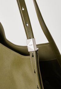 Sac fourre-tout en cuir vert olive avec une texture lisse, doté d'un accent de quincaillerie géométrique en argent et d'une bandoulière réglable avec perforations.