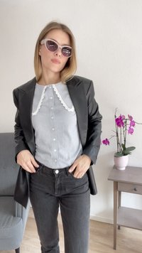 Blazer in ecopelle nera sopra una camicia azzurra con bottoni e colletto in pizzo bianco, abbinato a jeans scuri. Stanza con un'orchidea in vaso.