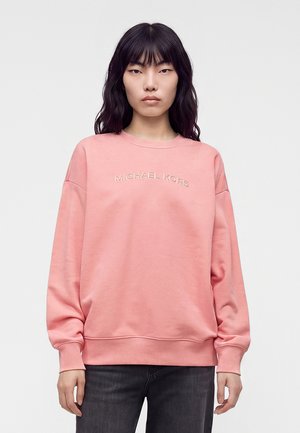 Kvinde med mørkt hår iført en lyserød Michael Kors sweatshirt og mørke bukser mod en ensfarvet lys baggrund, vendt fremad.