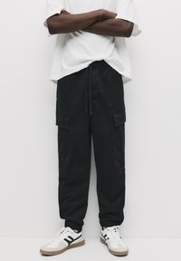 Zwarte cargobroek met elastische boorden, een trekkoord in de taille en meerdere voorzakken. Gecombineerd met witte sneakers met zwarte accenten.