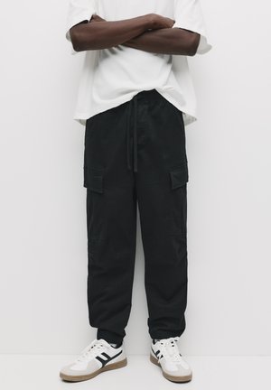 Zwarte cargobroek met elastische boorden, een trekkoord in de taille en meerdere voorzakken. Gecombineerd met witte sneakers met zwarte accenten.