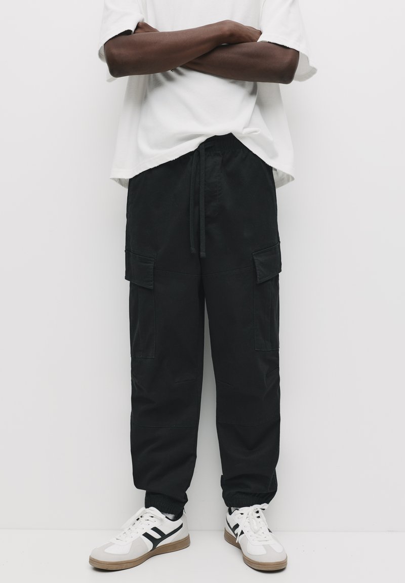 Zwarte cargobroek met elastische boorden, een trekkoord in de taille en meerdere voorzakken. Gecombineerd met witte sneakers met zwarte accenten.