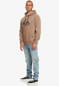 Brown hooded sweatshirt with a logo on the front, front pocket, paired with light blue jeans and black sneakers.