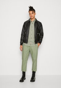 Chaqueta bomber de cuero negro sobre una camisa de botón verde, combinada con pantalones verde claro y botas negras de cordones, de pie contra un fondo blanco.