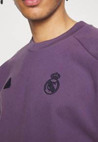 Sudadera púrpura con cuello acanalado, que presenta un logo bordado negro y un detalle de corona, junto a un emblema de marca negro más pequeño.