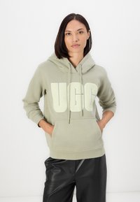 Gaiši zaļa kapuce, izgatavota no mīksta auduma, ar lielu, teksturētu UGG logo priekšpusē un kengura kabatu.