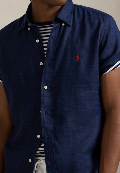 Mørkeblå linskjorte med korte ermer, knapp ned-krage og hvite knapper. Har en liten brodert rød logo og stripete undertrøye.