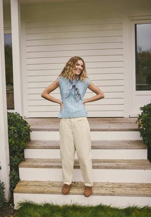 Top tricotat albastru deschis fără mâneci cu model texturat, combinat cu pantaloni largi crem. Modelul stă pe trepte de lemn, zâmbind.