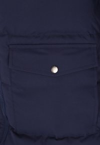 Sezione di giacca imbottita blu navy con una tasca anteriore dotata di bottone a pressione. Tessuto liscio con cuciture visibili lungo le cuciture.