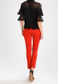 Sort bluse i blonder med udvidede ærmer, kombineret med figursyede røde bukser og sorte højhælede sko. Bagfra ses knapdetaljer og tekstur.