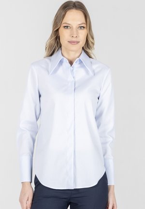 Kobieta ubrana w jasnoniebieską, długorękawową koszulę zapinaną na guziki z szerokim kołnierzem, stojąca na tle jednolitego szarego tła.