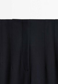Gros plan de la ceinture d'un pantalon noir montrant les lignes de couture verticales et le détail de la braguette sur un tissu lisse.