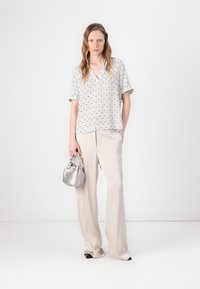 Blouse gris clair avec motif géométrique, associée à un pantalon large beige. Le modèle tient un petit sac à main argenté et se tient devant un fond uni.