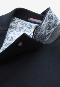 Col de veste bleu marine avec une doublure à motif géométrique bleu clair et blanc et une fermeture à bouton-pression argenté.