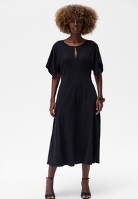 Schwarzes Midi-Kleid mit kurzen, weiten Ärmeln, einem Ausschnitt mit Schlüsselloch und einer fließenden, glatten Textur. Kombiniert mit schwarzen, hohen Sandalen.