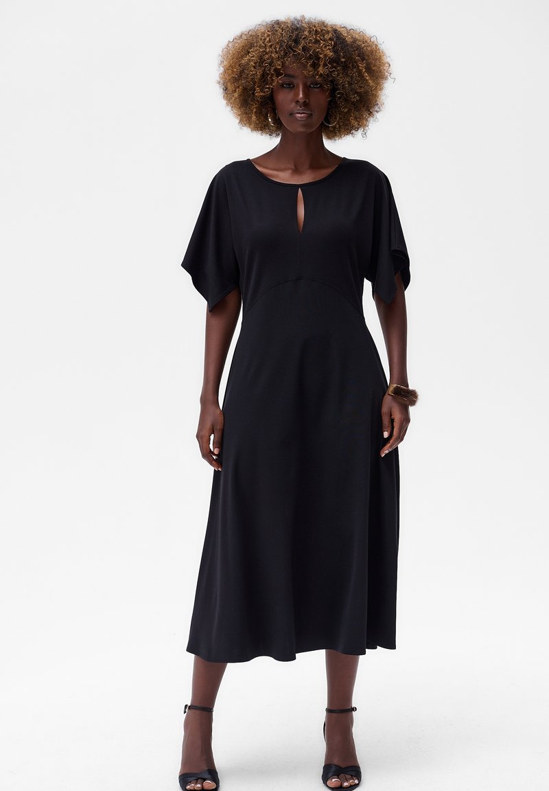 Schwarzes Midi-Kleid mit kurzen, weiten Ärmeln, einem Ausschnitt mit Schlüsselloch und einer fließenden, glatten Textur. Kombiniert mit schwarzen, hohen Sandalen.