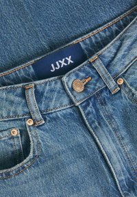 Jeans de denim azul claro con un botón y remaches visibles, costuras naranjas en contraste y una etiqueta azul oscuro que dice "JJXX" en la cinturilla.