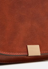 Brązowa skórzana torba o fakturze, z narożnikiem i prostokątnym złotym zapięciem oraz kontrastującym czarnym przeszyciem wzdłuż krawędzi.