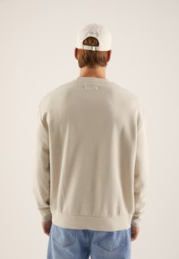 Light beige sweatshirt with ribbed hem and cuffs, featuring a soft texture. Worn with blue denim jeans and a white cap at the back.