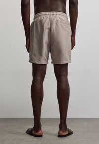 Homme debout pieds nus portant un short beige et des sandales noires sur un fond uni gris clair.