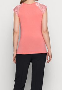 Korálově růžový přiléhavý top s krajkovými krátkými rukávy, hladká tkanina, kulatý výstřih a rovný lem. Kombinováno s tmavými, volně střiženými kalhotami.