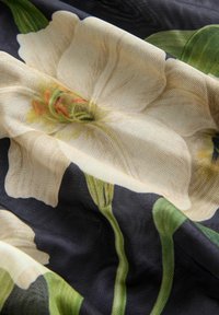 Tissu avec un grand motif de fleur blanche et des feuilles vertes sur un fond sombre, montrant la texture et les plis du matériau.