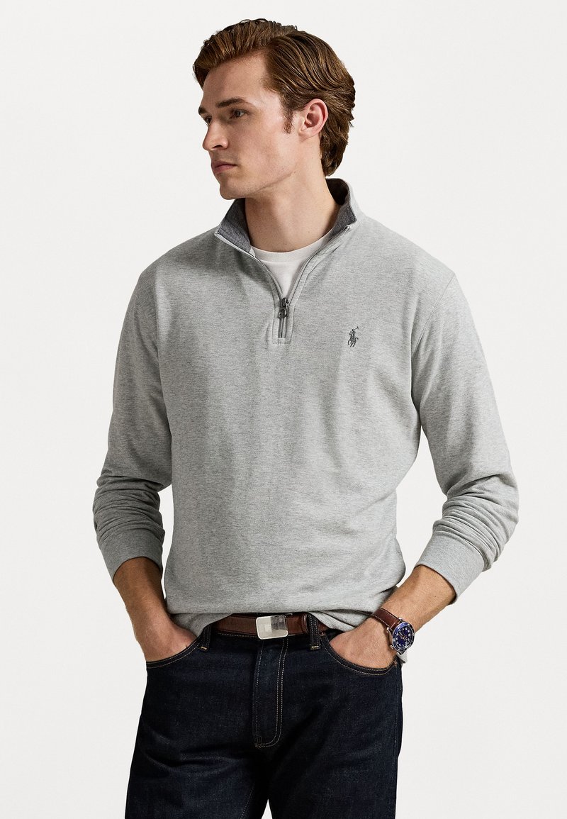 Sudadera de felpa de algodón gris con media cremallera y cuello y puños acanalados. Presenta un pequeño logotipo bordado en el pecho. Jeans de mezclilla oscura visibles.