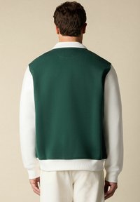 Man in witte lange mouwen shirt en crèmekleurige broek met een donkergroene mouwloze vest over het shirt, staand tegen een effen beige achtergrond.