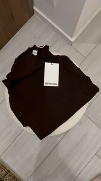 Barna bordázott pulóver, egy kerek fehér asztalon elhelyezve. A címkén a "BERSHKA" felirat olvasható. A szövet puha, texturált felületűnek tűnik.