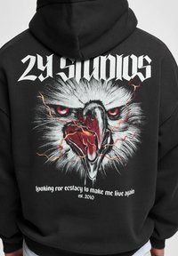 Mustmust särav must kapuutsiga, mille seljal on detailne kotka graafika, punaste silmade ja välkudega. Tekst rõhutab "29 STUDIOS" ja moto.