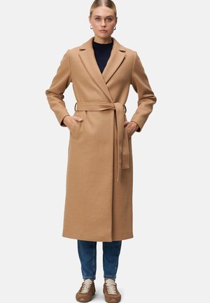 Manteau enveloppant couleur camel en tissu doux, doté d'une taille ceinturée, d'un col cranté, de deux poches latérales, assorti à un jean bleu et des baskets.