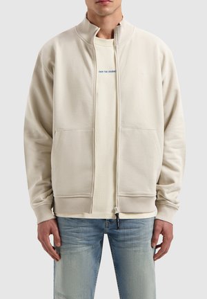 Beige zip-up sweatshirt with high collar, ribbed cuffs, two front pockets, and a subtle logo on the chest. Worn over a light-colored shirt.