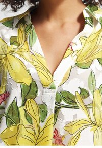 Blouse fleurie avec des lys jaunes et des feuilles vertes sur fond blanc. Encolure en V et tissu doux et texturé avec des motifs subtils.