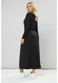 Abito maxi a coste nere con maniche lunghe, silhouette aderente, scollatura rotonda e fessure laterali, abbinato a sneakers bianche.