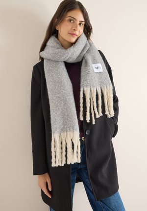 Écharpe grise et crème avec des détails à franges, fabriquée en matériau doux ; portée sur un manteau sombre sur un fond neutre. Étiquette visible.